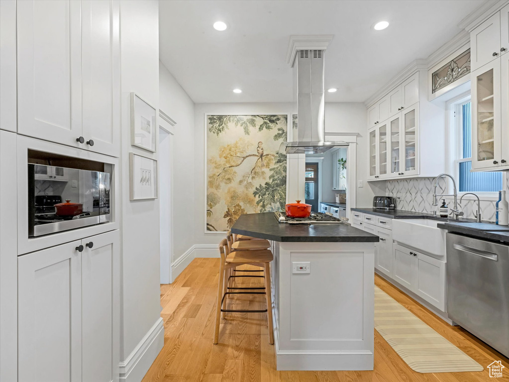 Kitchen featuring stainless steel appliances, island range hood, dark countertops, light wood-style floors, and white cabinetry