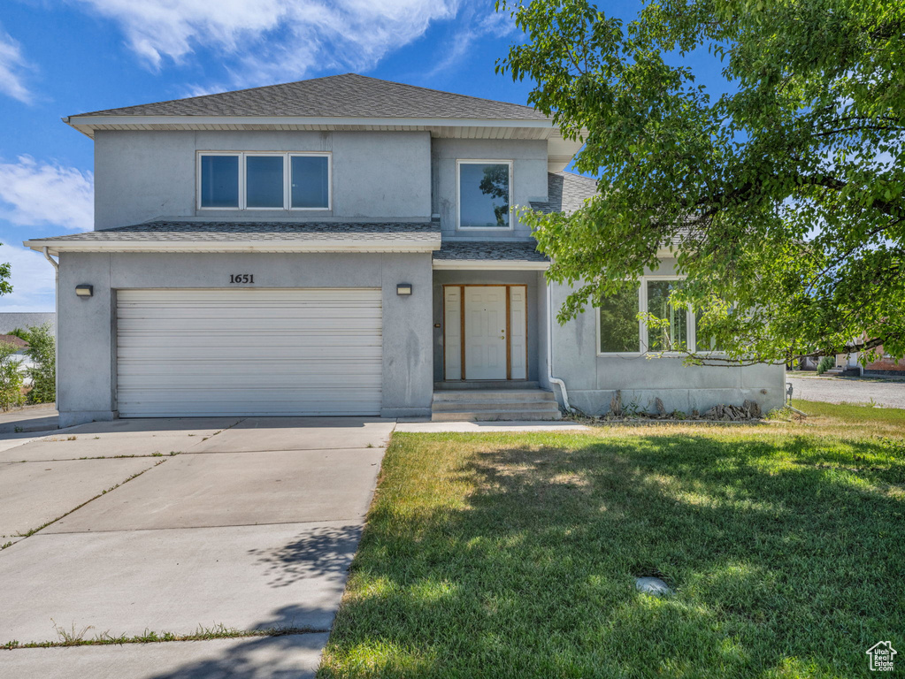 View of front facade with stucco siding, an attached garage, a front yard, driveway, and a shingled roof