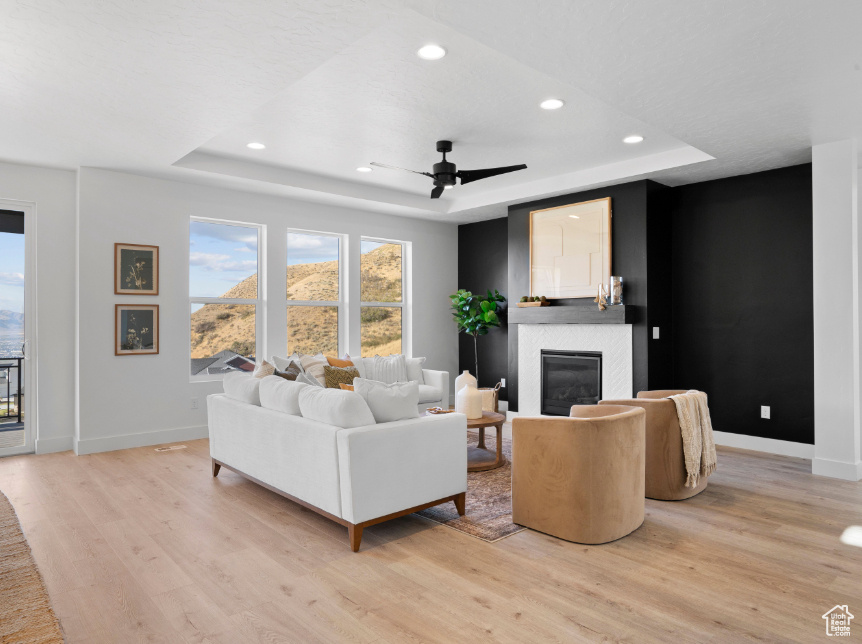 Living area with a raised ceiling, light wood-style flooring, a glass covered fireplace, recessed lighting, and a ceiling fan