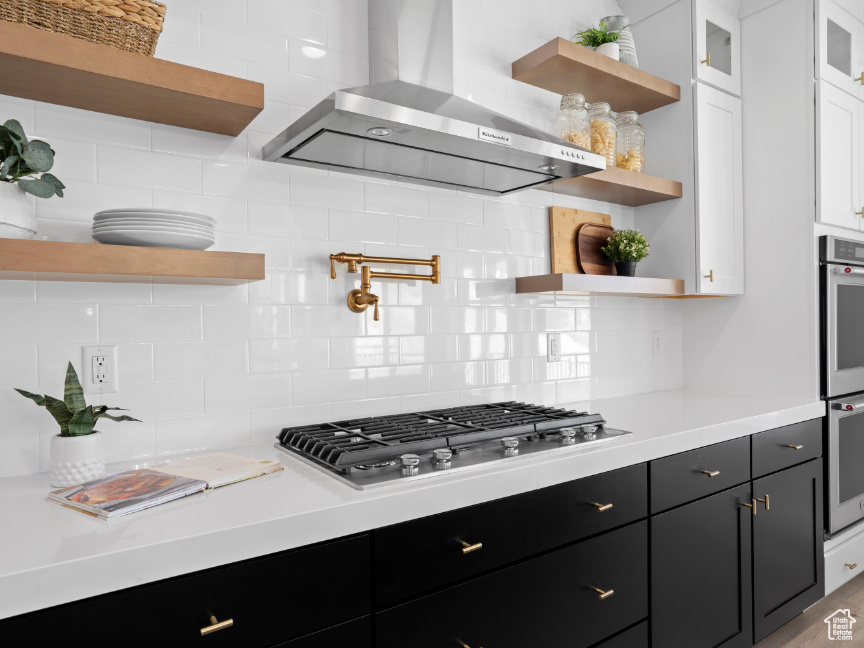 Kitchen with open shelves, wall chimney exhaust hood, stainless steel gas stovetop, white cabinets, and light countertops