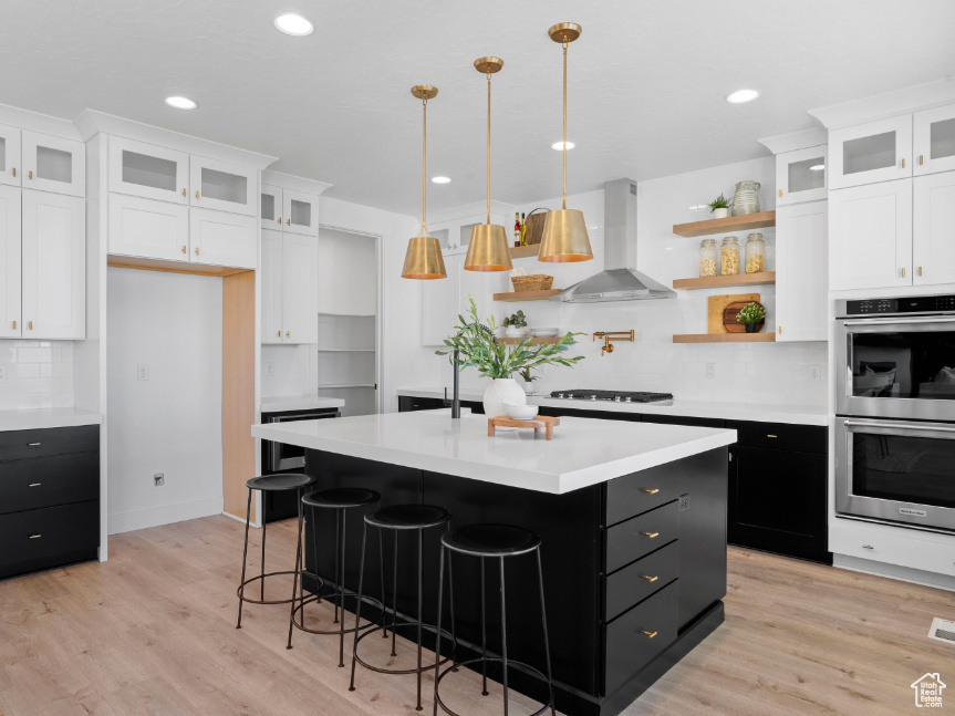 Kitchen featuring wall chimney range hood, dark cabinets, stainless steel double oven, white cabinetry, and light countertops