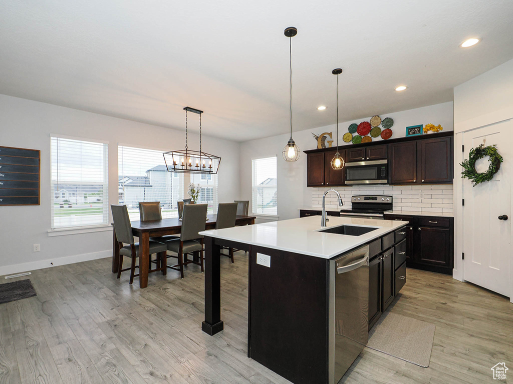 Kitchen with appliances with stainless steel finishes, light wood-type flooring, tasteful backsplash, light countertops, and an island with sink