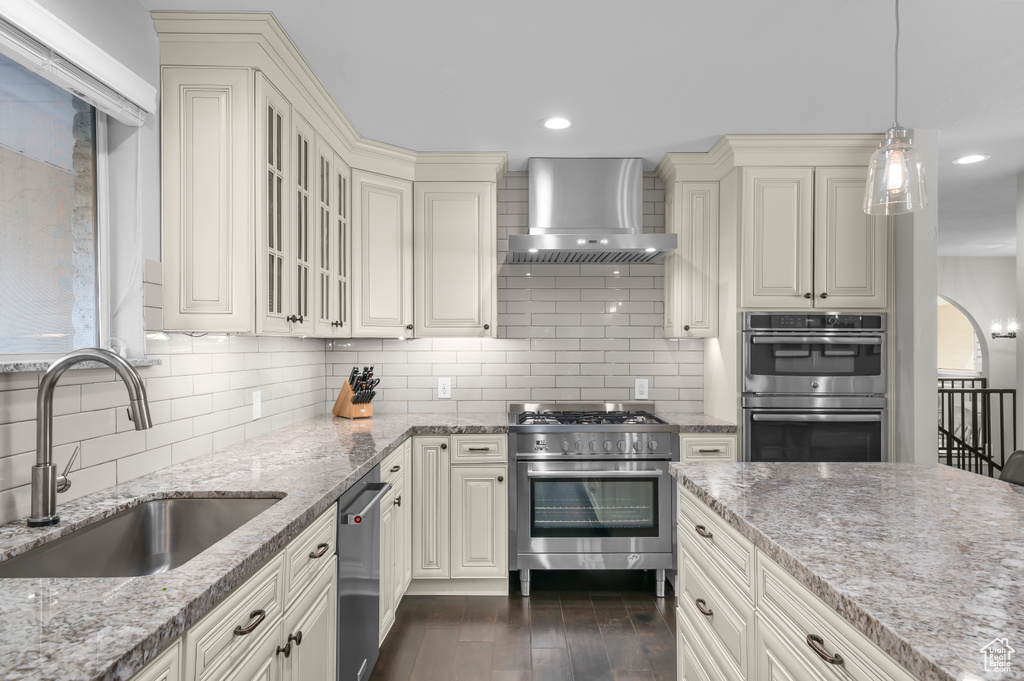 Kitchen with wall chimney range hood, appliances with stainless steel finishes, cream cabinetry, light stone counters, and decorative backsplash