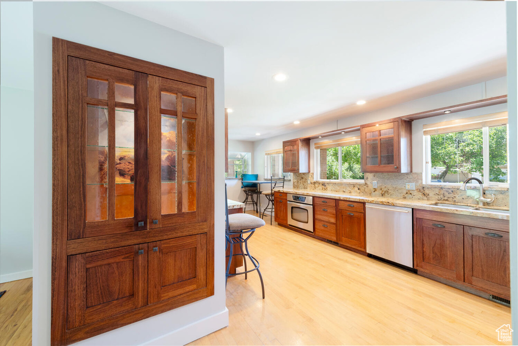 Kitchen featuring appliances with stainless steel finishes, brown cabinetry, light wood-style floors, decorative backsplash, and glass insert cabinets