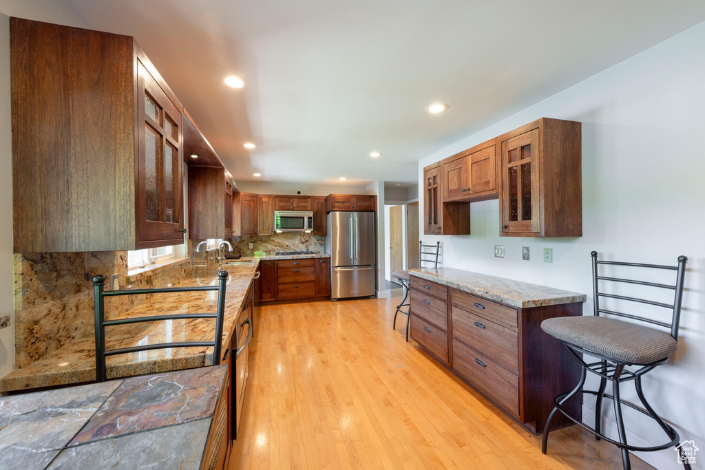 Kitchen featuring stainless steel appliances, glass insert cabinets, tasteful backsplash, light wood-type flooring, and light stone counters