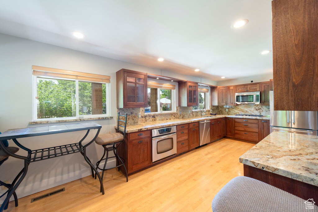 Kitchen featuring appliances with stainless steel finishes, tasteful backsplash, light stone countertops, light wood-type flooring, and glass insert cabinets