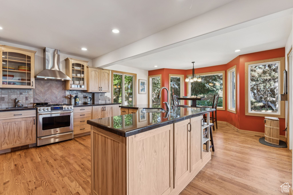 Kitchen featuring wall chimney range hood, stainless steel gas range, light brown cabinets, dark countertops, and recessed lighting