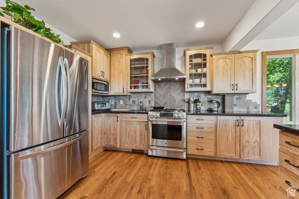 Kitchen featuring appliances with stainless steel finishes, wall chimney range hood, light brown cabinetry, dark countertops, and recessed lighting