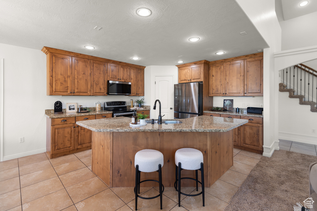 Kitchen featuring stainless steel appliances, brown cabinetry, light tile patterned flooring, an island with sink, and recessed lighting