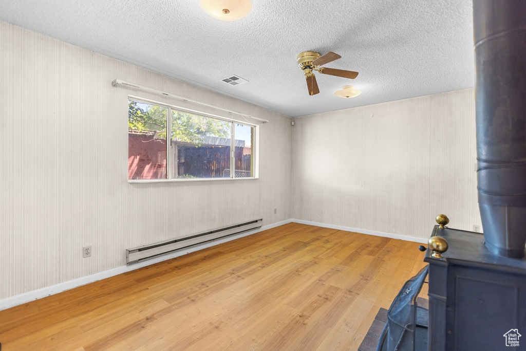 Empty room with baseboard heating, wood finished floors, a ceiling fan, a textured ceiling, and a wood stove