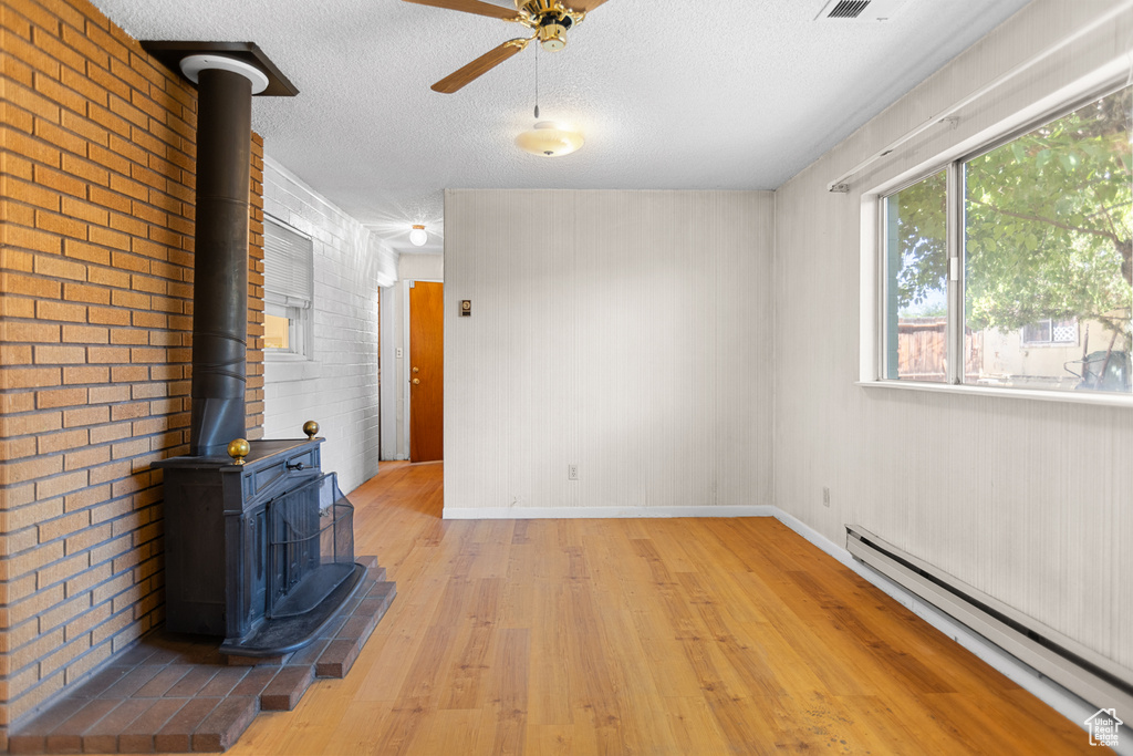 Unfurnished living room with a baseboard heating unit, a wood stove, light wood finished floors, a ceiling fan, and a textured ceiling