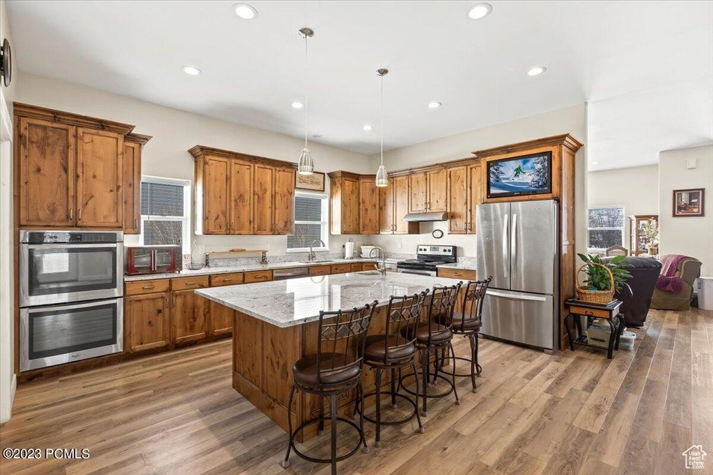 Kitchen featuring stainless steel appliances, brown cabinetry, wood finished floors, an island with sink, and under cabinet range hood