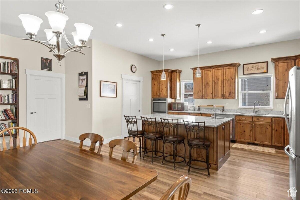 Kitchen with appliances with stainless steel finishes, brown cabinetry, an island with sink, light wood-style flooring, and a chandelier