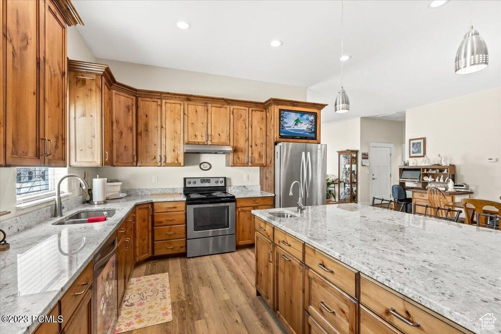 Kitchen featuring stainless steel appliances, under cabinet range hood, brown cabinets, light wood-type flooring, and light stone countertops