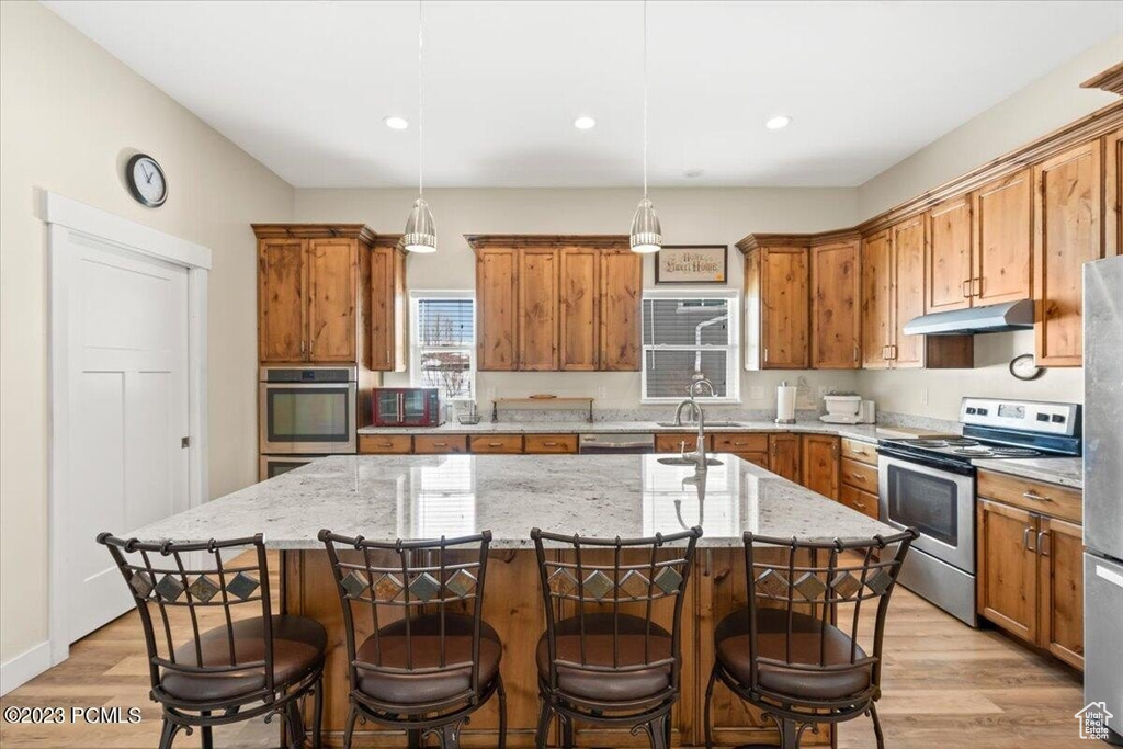 Kitchen featuring stainless steel appliances, brown cabinets, under cabinet range hood, light wood-style floors, and recessed lighting
