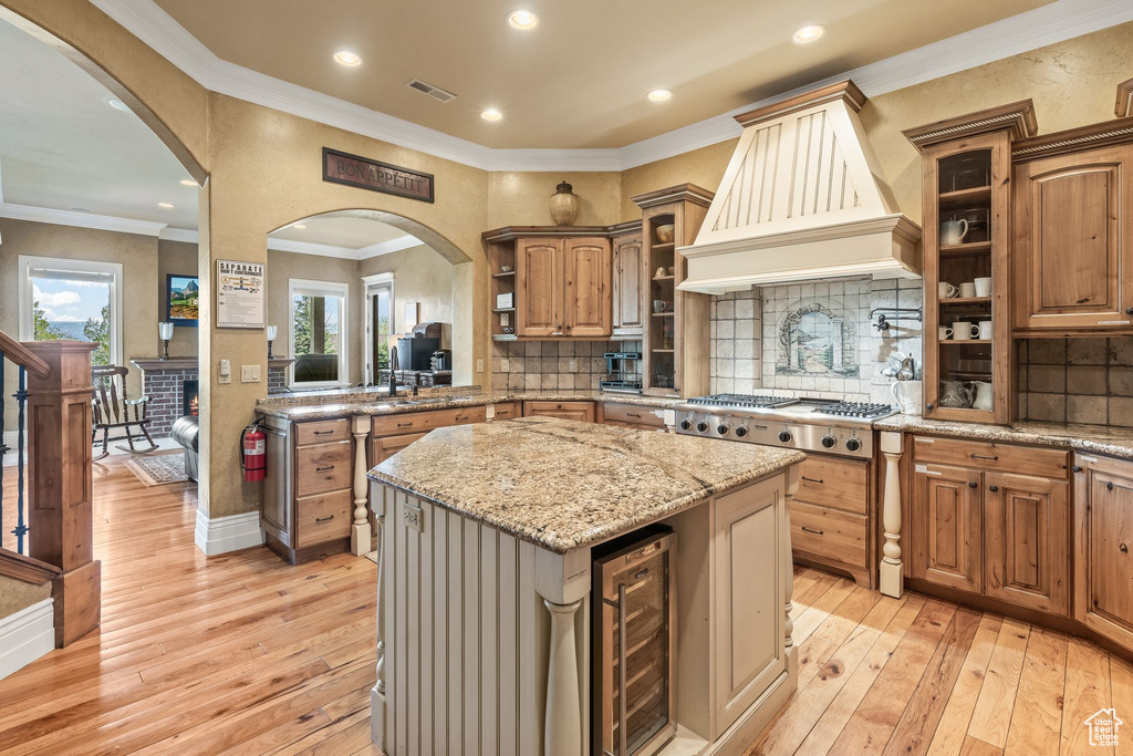 Kitchen with beverage cooler, custom exhaust hood, stainless steel gas stovetop, a kitchen island, and light wood-style floors