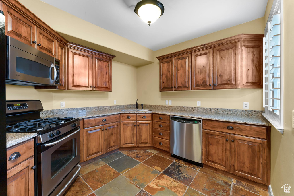 Kitchen featuring stainless steel appliances, stone tile floors, brown cabinetry, and light stone countertops