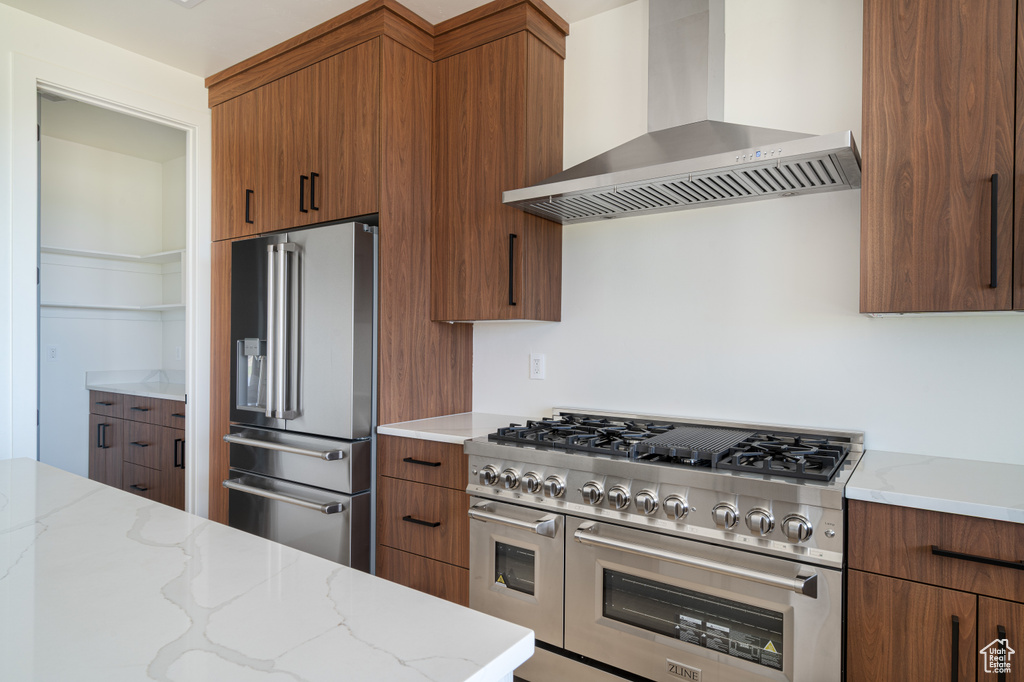 Kitchen featuring wall chimney range hood, premium appliances, light stone countertops, and brown cabinetry