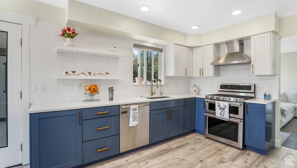 Kitchen with wall chimney range hood, stainless steel appliances, blue cabinetry, light wood-type flooring, and light countertops