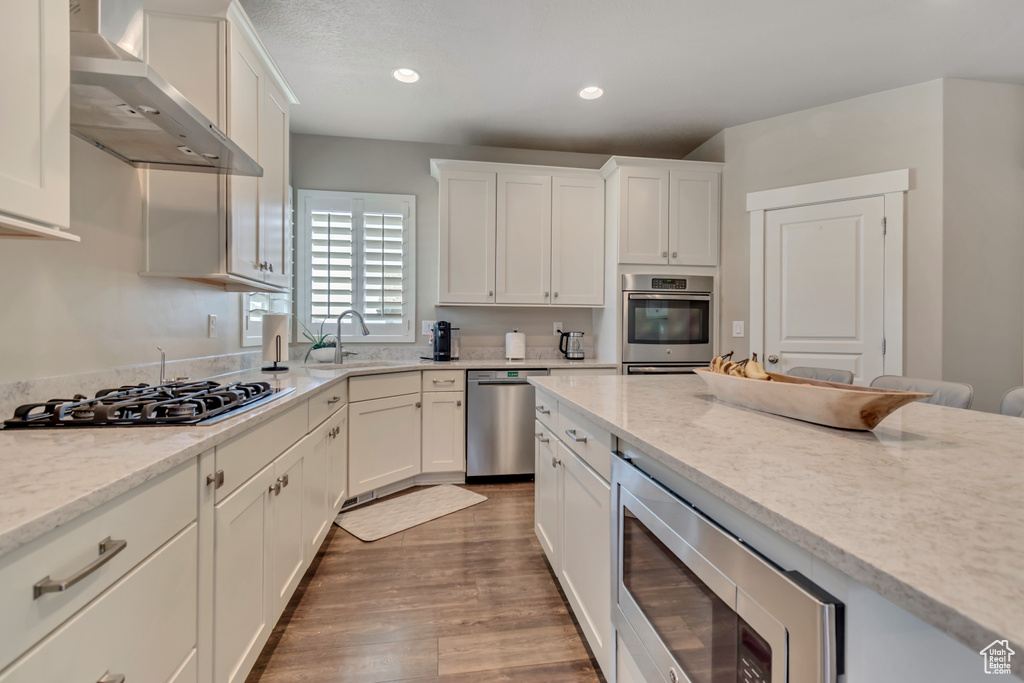 Kitchen with stainless steel appliances, wall chimney exhaust hood, white cabinets, wood finished floors, and recessed lighting