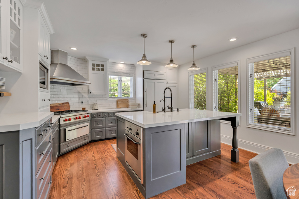 Kitchen with gray cabinetry, wall chimney exhaust hood, light countertops, built in appliances, and tasteful backsplash