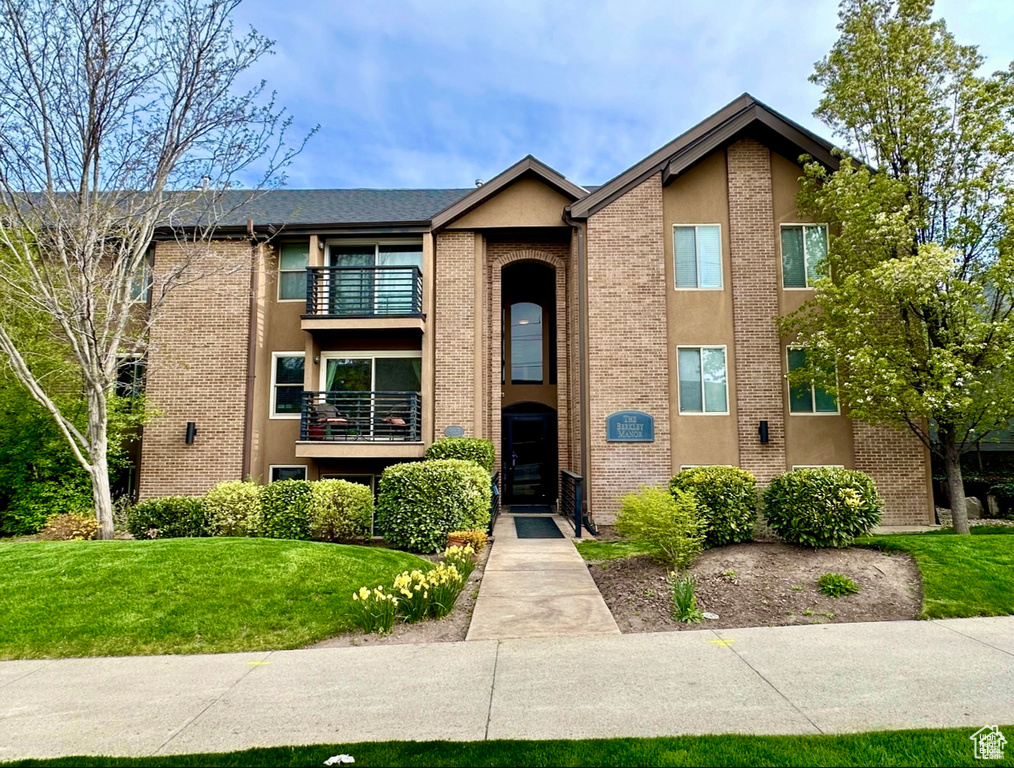View of front facade featuring a balcony, brick siding, a front yard, and stucco siding