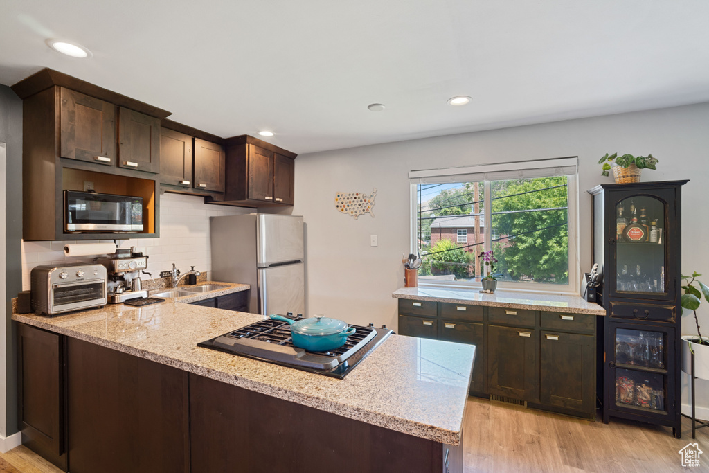 Kitchen featuring stainless steel appliances, dark brown cabinetry, light wood-type flooring, light stone countertops, and tasteful backsplash