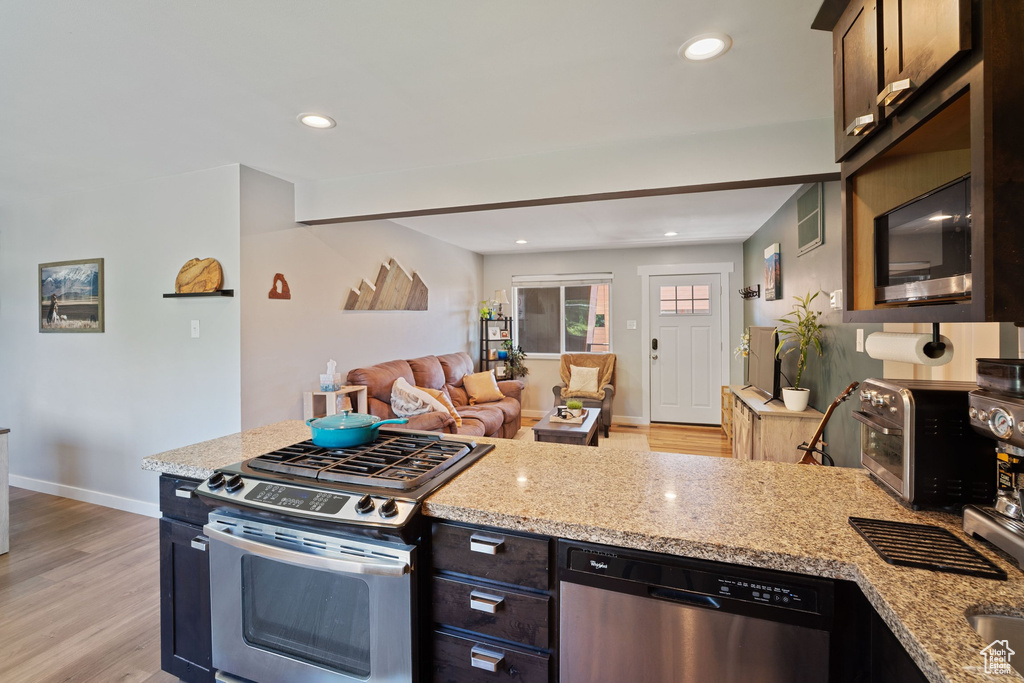 Kitchen with stainless steel appliances, light wood-type flooring, open floor plan, light stone counters, and dark brown cabinets