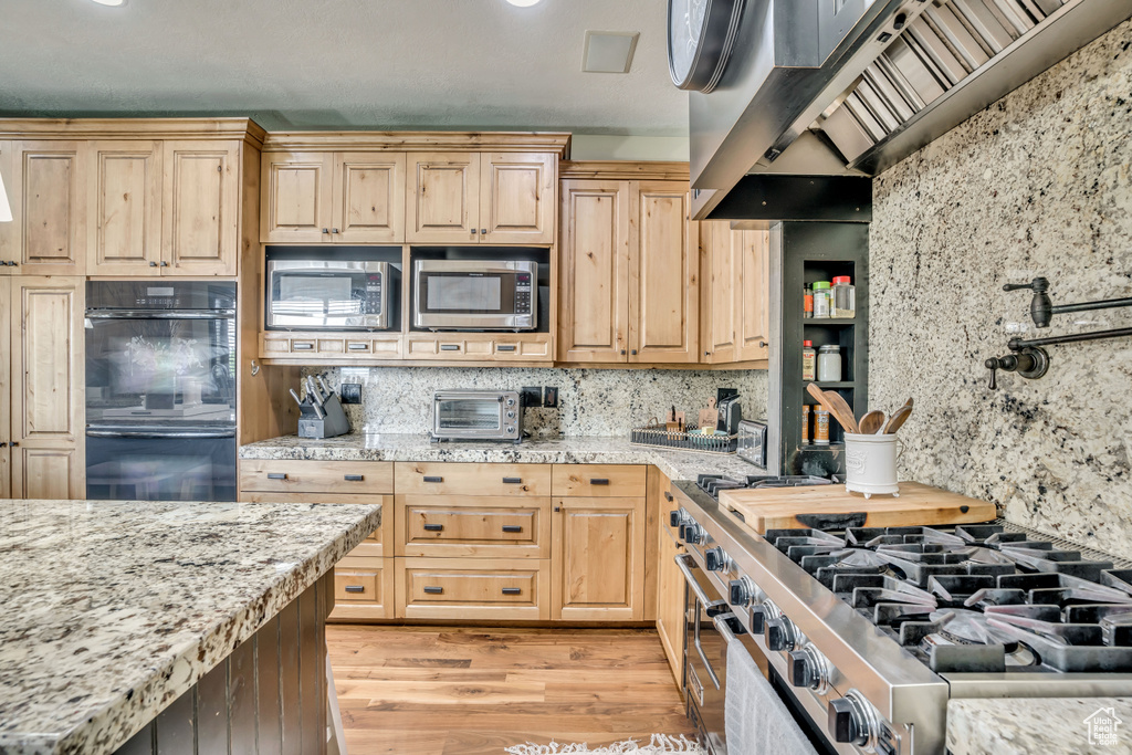 Kitchen featuring appliances with stainless steel finishes, custom range hood, light wood finished floors, backsplash, and light stone countertops
