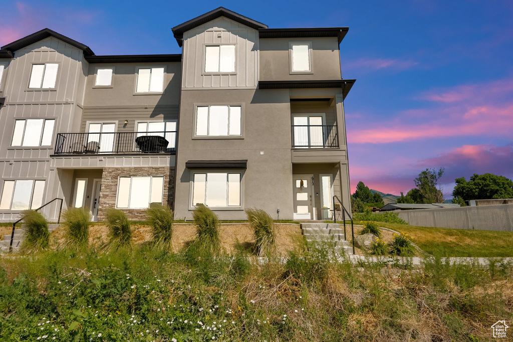 Back of property at dusk featuring board and batten siding, a balcony, stucco siding, and stone siding