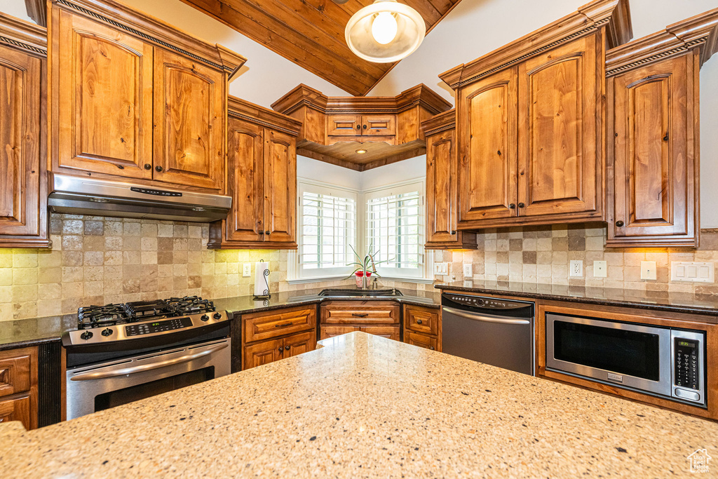 Kitchen with stainless steel appliances, under cabinet range hood, dark stone countertops, brown cabinets, and tasteful backsplash
