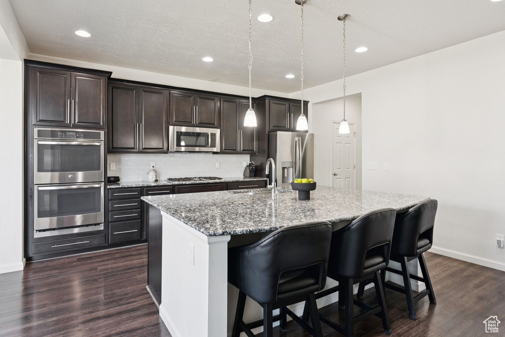 Kitchen with appliances with stainless steel finishes, an island with sink, dark wood-style floors, tasteful backsplash, and dark brown cabinets