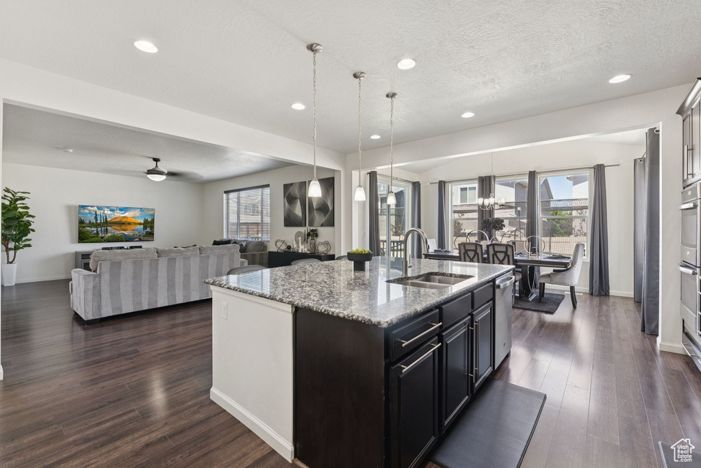 Kitchen with dark wood-style flooring, an island with sink, hanging light fixtures, recessed lighting, and light stone countertops