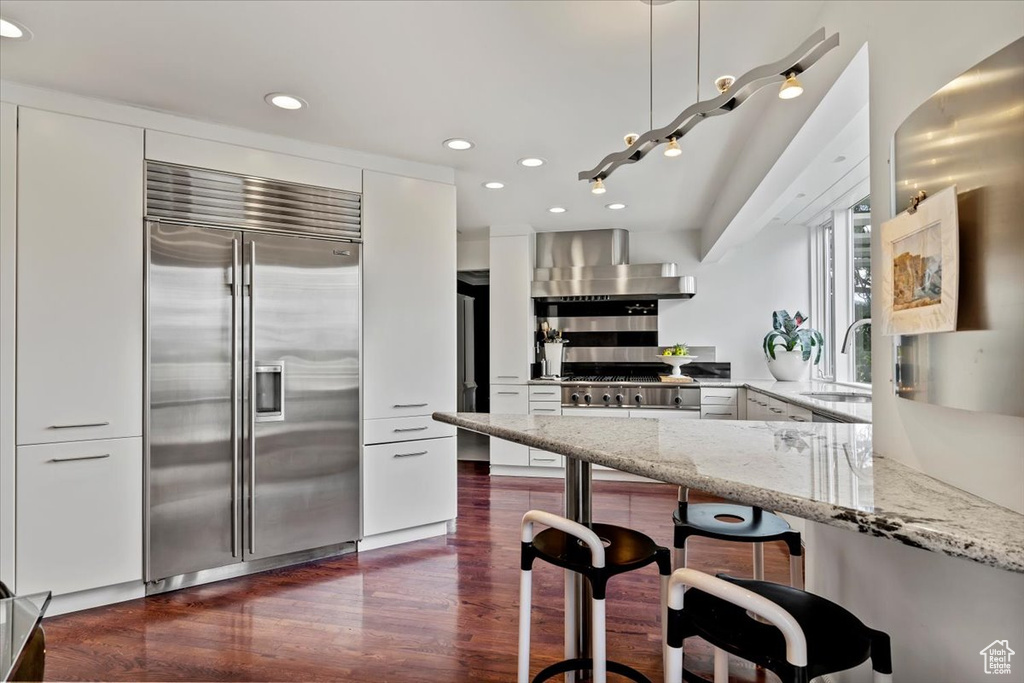 Kitchen featuring built in refrigerator, wall chimney exhaust hood, light stone countertops, dark wood-type flooring, and recessed lighting