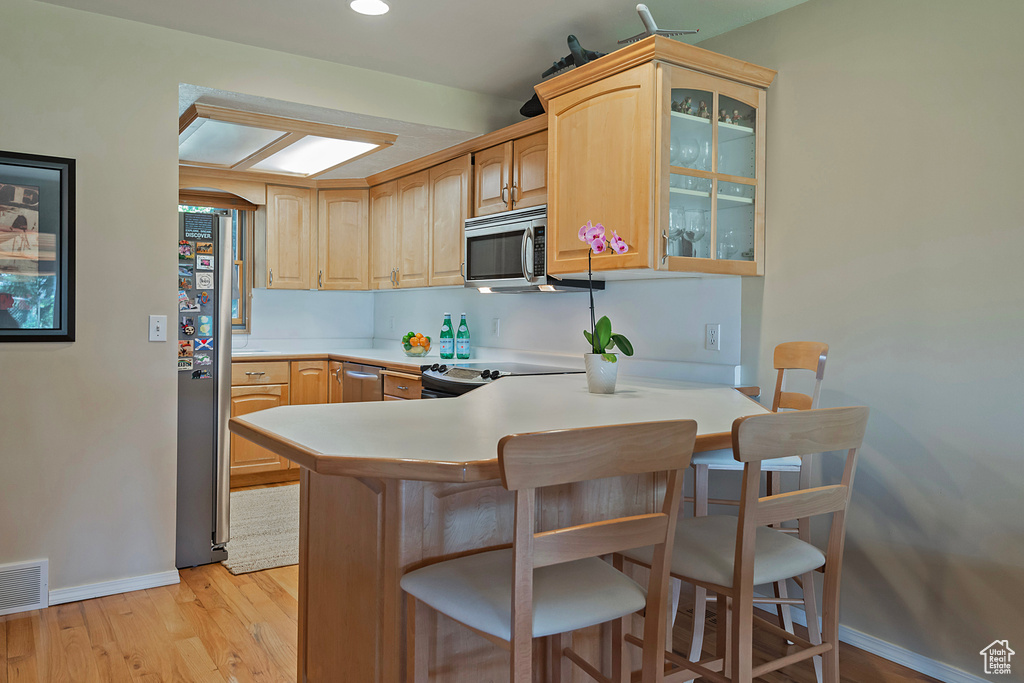 Kitchen with appliances with stainless steel finishes, glass insert cabinets, a breakfast bar area, light wood-style flooring, and a peninsula