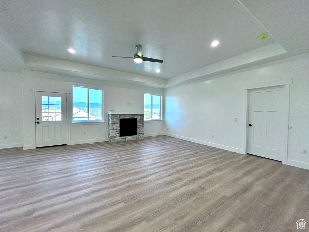 Unfurnished living room with a tray ceiling, ceiling fan, light wood-style flooring, a stone fireplace, and recessed lighting