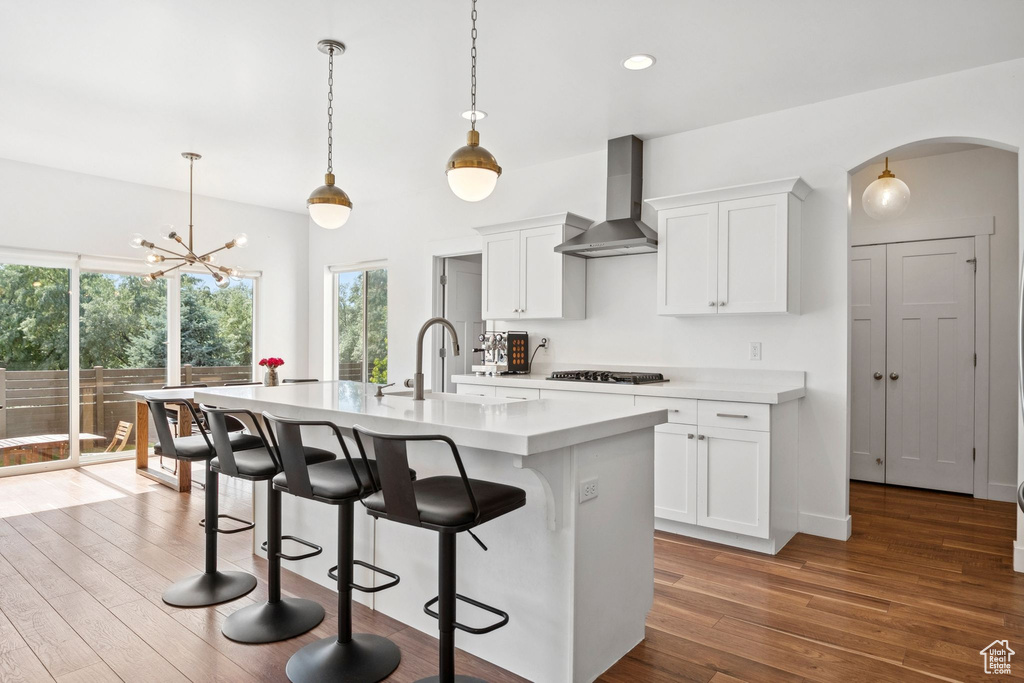 Kitchen featuring wall chimney exhaust hood, gas stovetop, arched walkways, light countertops, and a center island with sink