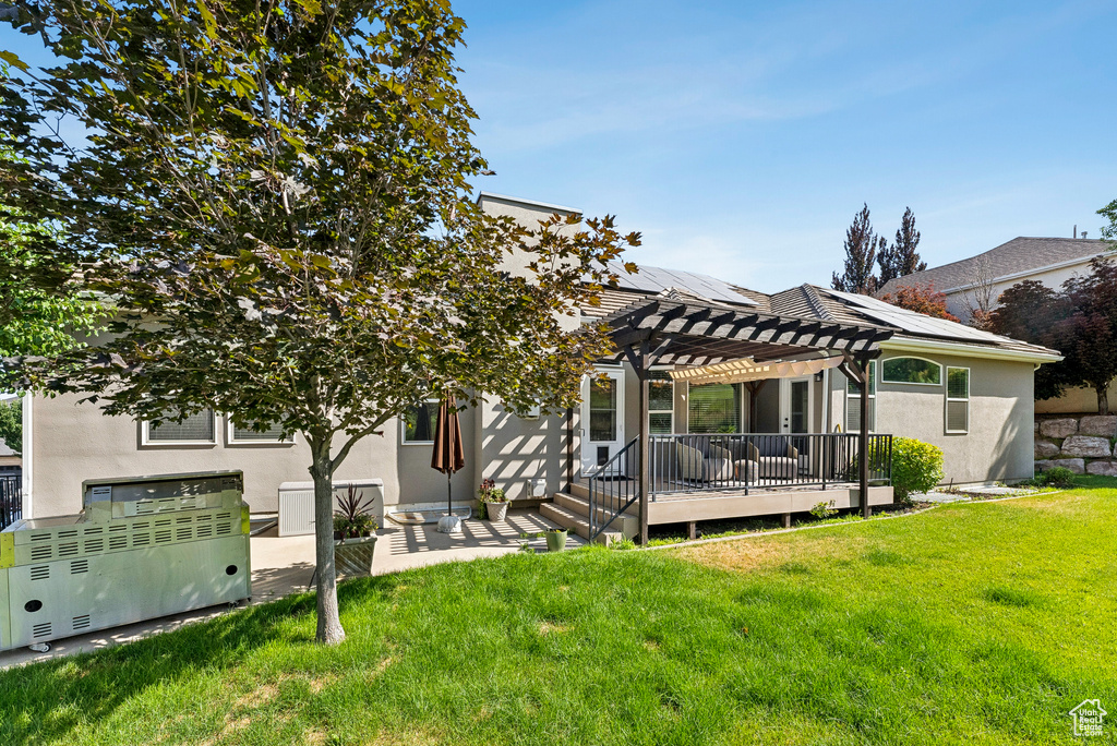 Rear view of property with a pergola, stucco siding, roof mounted solar panels, a lawn, and a deck