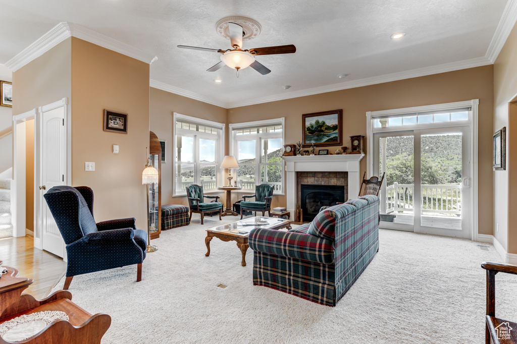 Carpeted living room featuring ceiling fan, a tiled fireplace, healthy amount of natural light, crown molding, and stairs