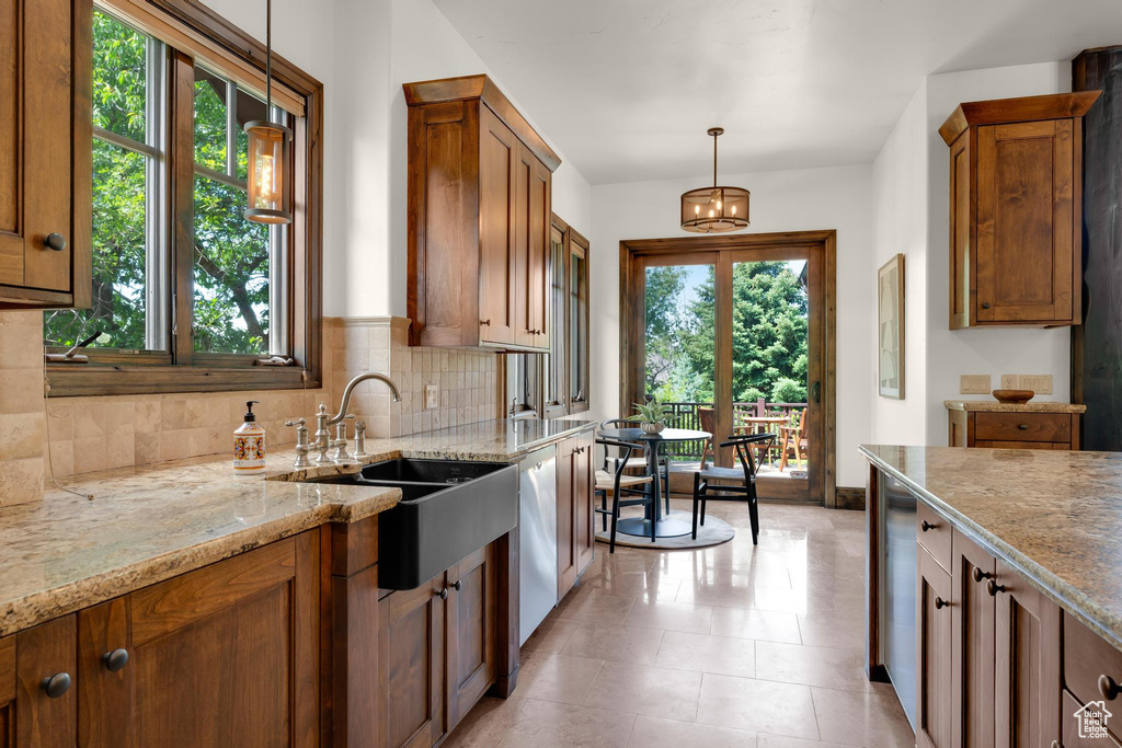 Kitchen with brown cabinetry, backsplash, light stone countertops, hanging light fixtures, and white dishwasher
