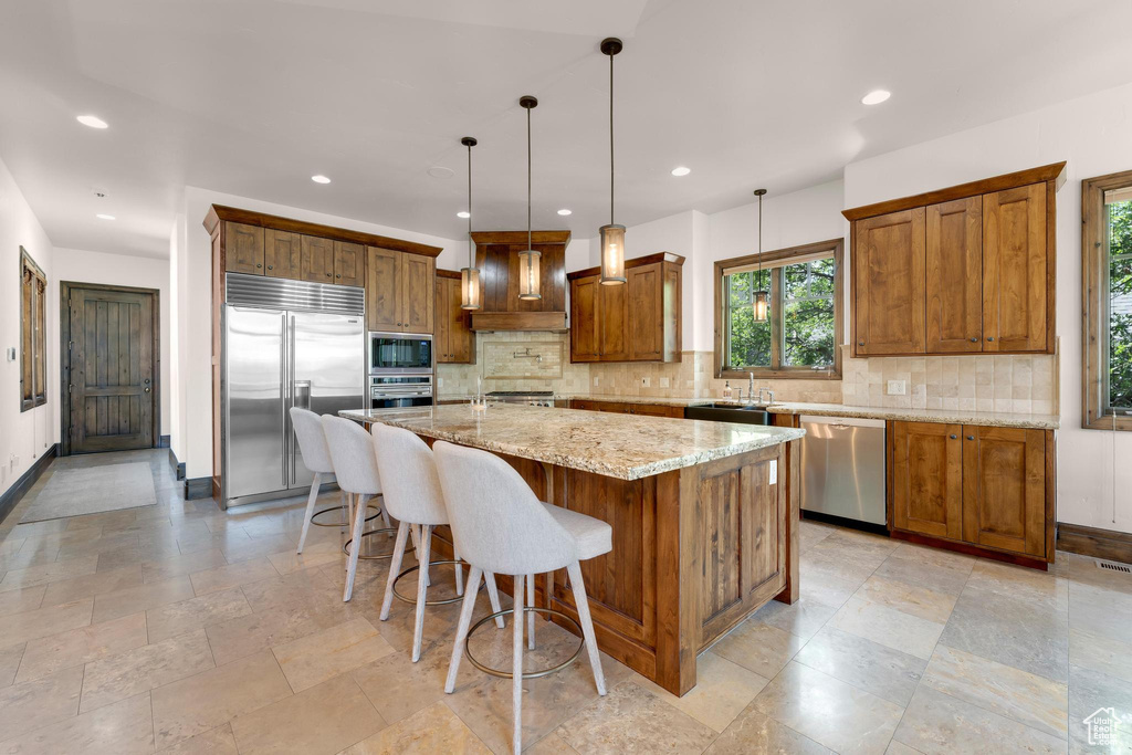 Kitchen with built in appliances, brown cabinetry, decorative backsplash, an island with sink, and light stone counters