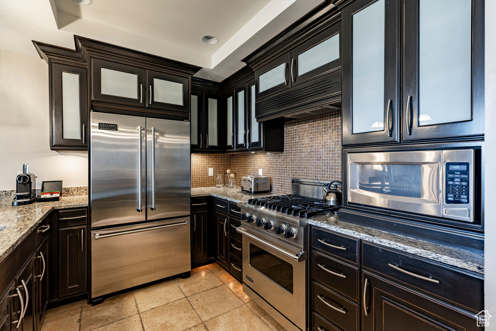 Kitchen featuring built in appliances, backsplash, light stone counters, and glass insert cabinets
