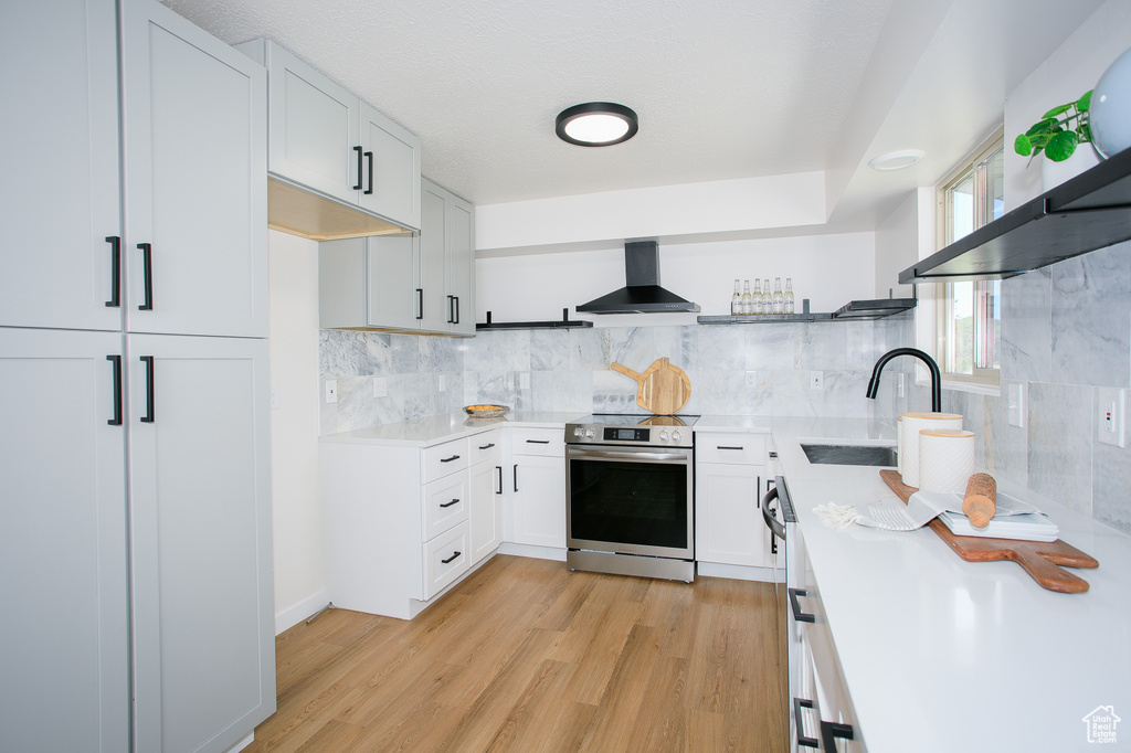 Kitchen with stainless steel electric stove, wall chimney range hood, light countertops, light wood-type flooring, and open shelves