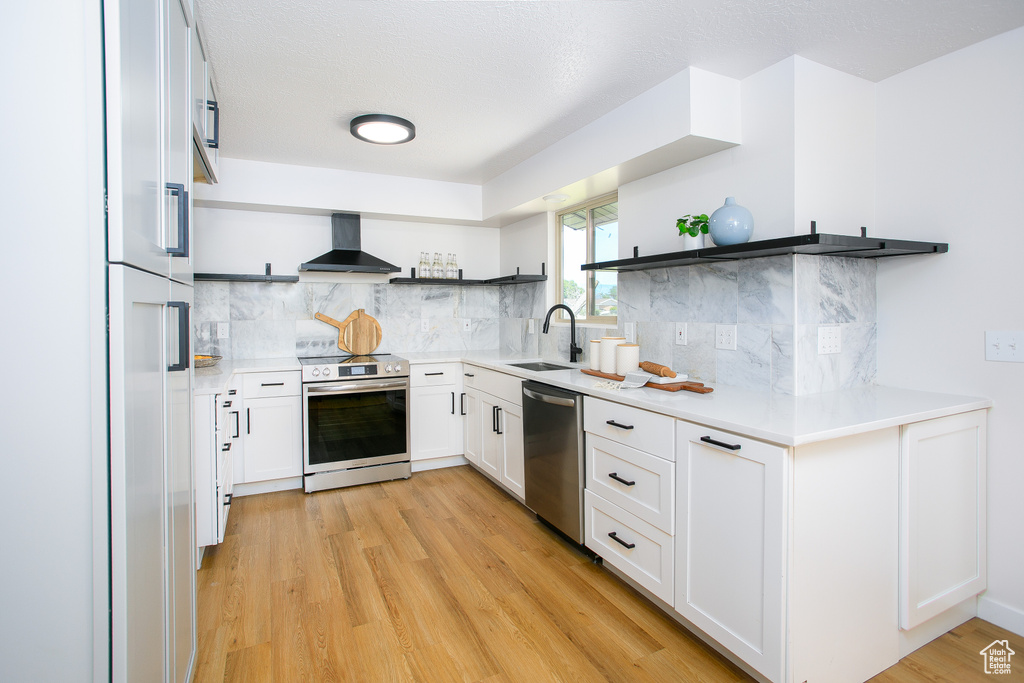 Kitchen featuring open shelves, appliances with stainless steel finishes, wall chimney range hood, light wood-style floors, and tasteful backsplash