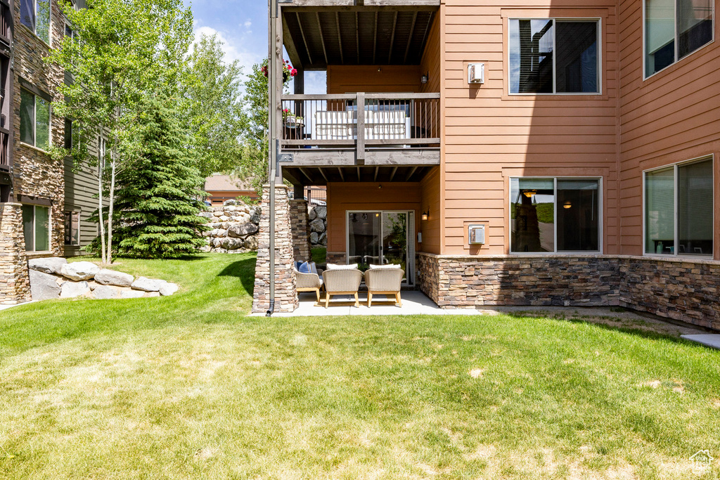 Back of house featuring a balcony, stone siding, a lawn, and a patio