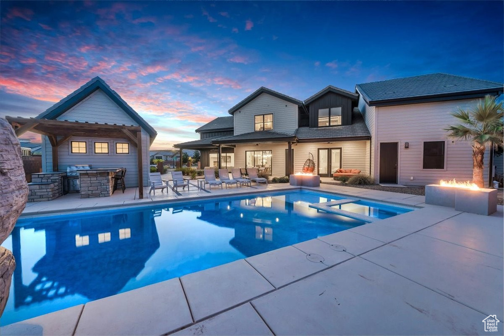 Pool at dusk with a fire pit, a patio area, exterior bar, exterior kitchen, and a pool with connected hot tub