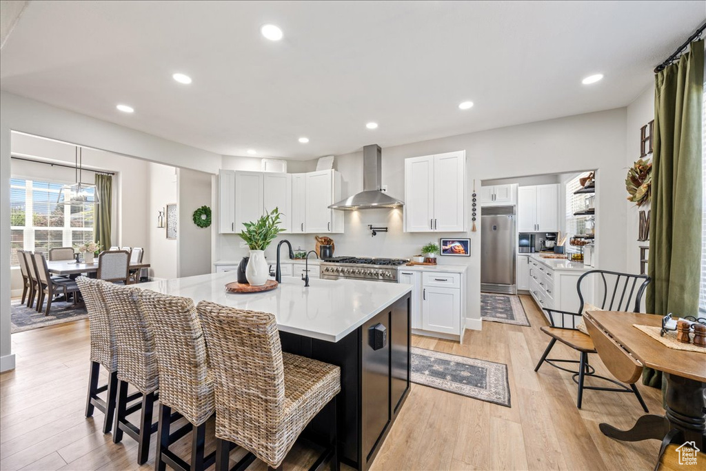 Kitchen featuring wall chimney range hood, built in fridge, light countertops, light wood finished floors, and a center island with sink