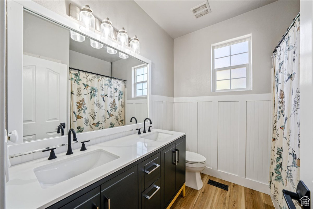 Full bath featuring a wainscoted wall, double vanity, wood finished floors, plenty of natural light, and a decorative wall