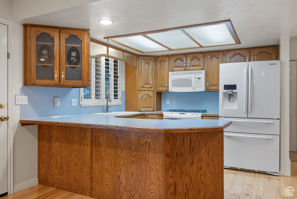 Kitchen with white appliances, brown cabinetry, glass insert cabinets, light wood-style flooring, and a textured ceiling