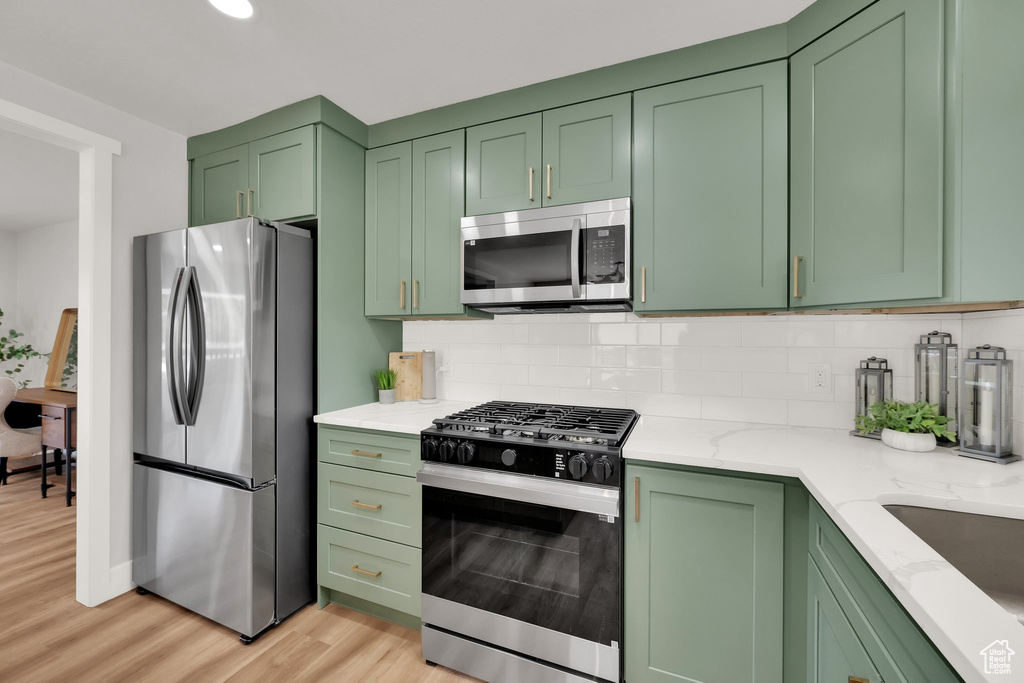 Kitchen with stainless steel appliances, green cabinets, light wood-style floors, decorative backsplash, and light stone countertops